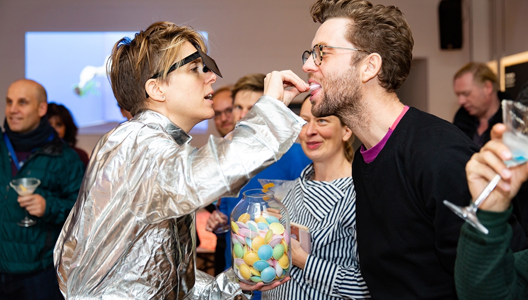 EMILIE BALTZ FEEDS MAN PINK OBJECT WITH AUDIENCE WATCHING IN EAT TECH KITCHEN