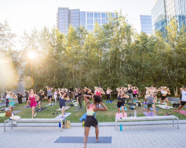 PEOPLE PRACTICING YOGA IN UNISON OUTSIDE AT BOSTON SEAPORT SURROUNDED BY TREES AND BUILDINGS