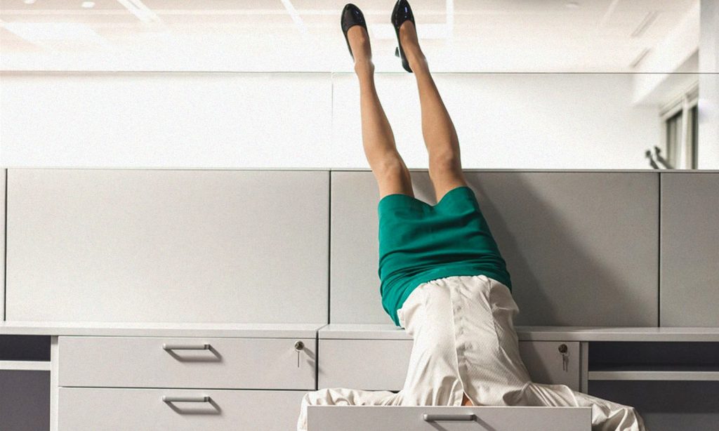WOMAN IN AN OFFICE CUBICLE LYING UPSIDE DOWN WITH HER HEAD IN DESK DRAWER AND FEET UP IN THE AIR