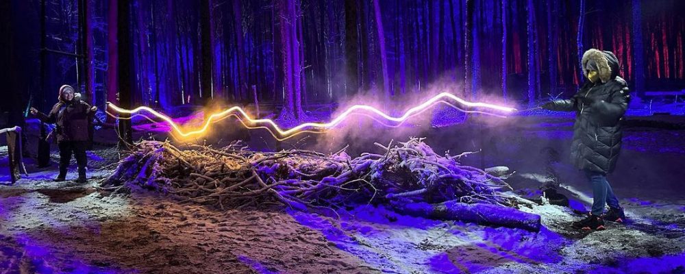 TWO PEOPLE HOLD WANDS IN FOREST WITH NEON LIGHT CONNECTING THEM IN HARRY POTTER: A FORBIDDEN FOREST EXPERIENCE