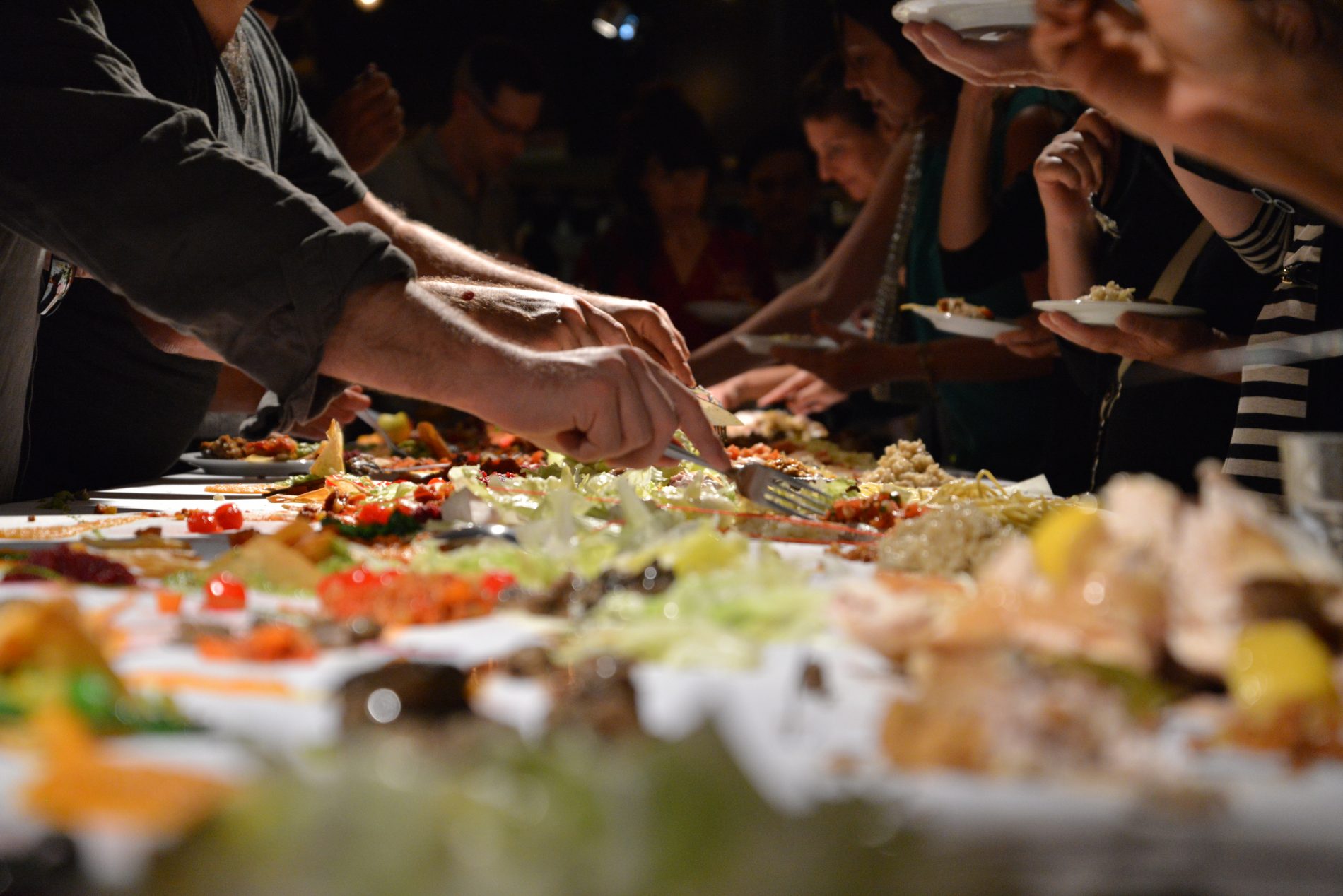 CLOSE UP OF HANDS AND UTENSILS GRABBING FOOD THAT COVER A TABLE IN SEMI-ANNUAL DINNER CLUB CALLED IN THE MOUTH