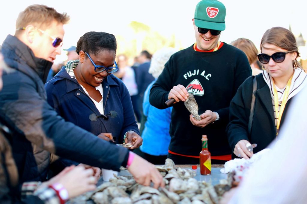 March XP: Group of people enjoying an outdoor food festival, shucking fresh oysters and sharing smiles.