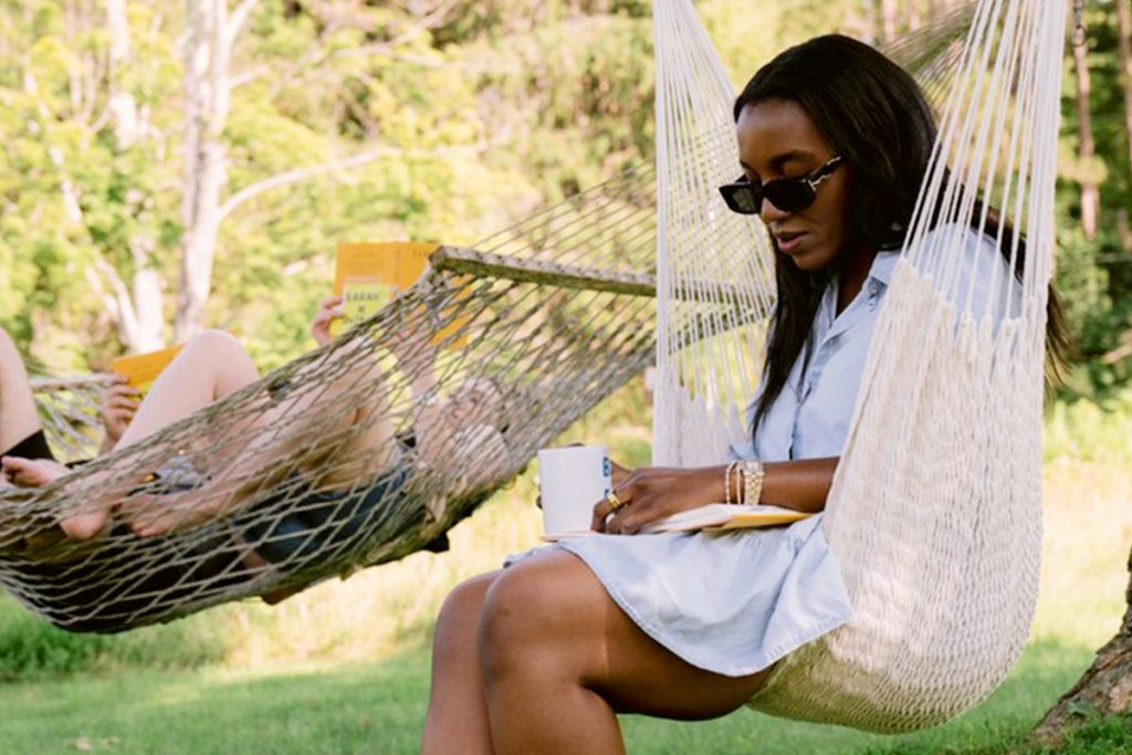March XP: Woman relaxing in a hammock outdoors, reading a book and sipping a drink in a peaceful, natural setting.